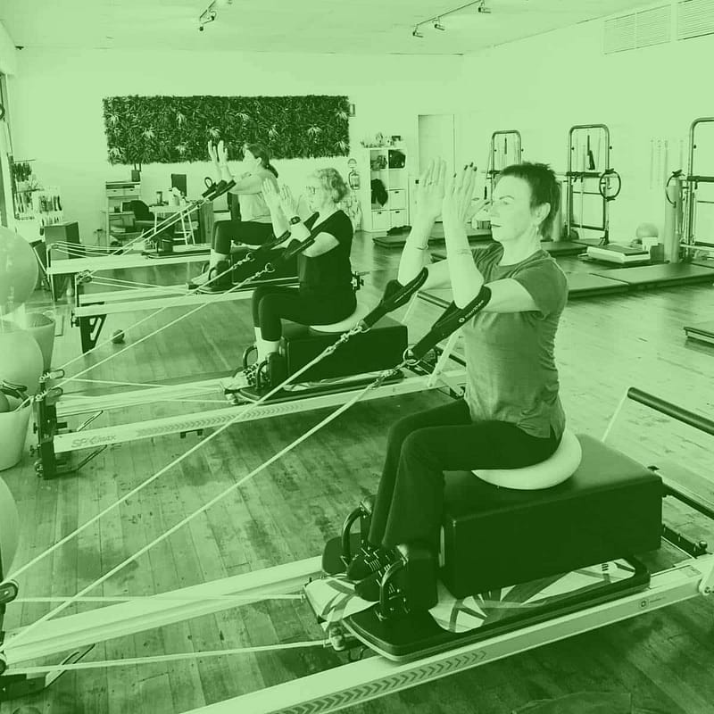 Our Classes 14 women doing seated arm exercises in a reformer pilates class